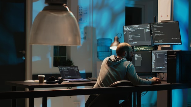 IT system engineer coding interface on multiple monitors to program new server system late at night. Working after hours to develop source code on terminal window, AI html language.