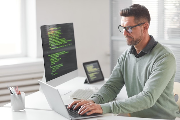 Mature computer programmer programming and coding new technologies while typing on laptop at the table at office