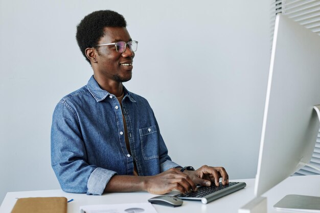 Minimal portrait of smiling black man using pc at workplace in office against white wall copy space