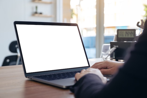 Mockup image of hands using and typing on laptop with blank white desktop screen on wooden table in office