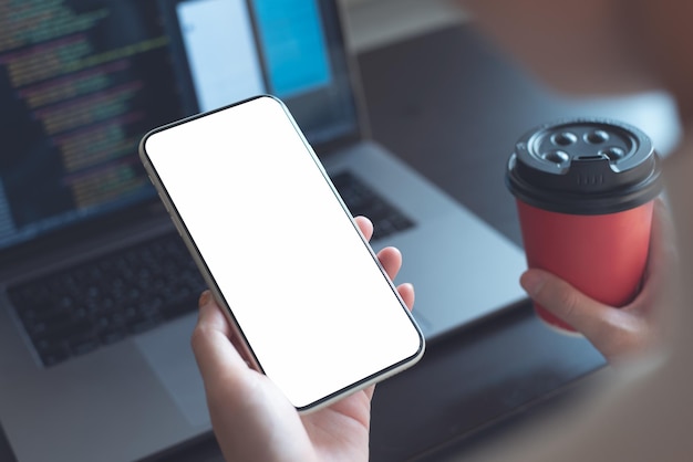 Mockup image woman holding mobile phone with blank screen template during working on laptop