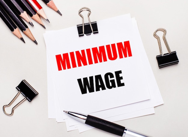 On a light background black pencils black paper clips a pen and a sheet of white paper with the text MINIMUM WAGE