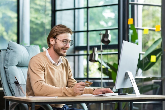 Online transactions with a credit card a smiling young man sits in the office at the table looks at