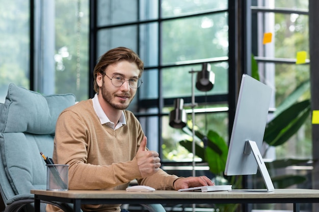 Portrait of a young man in glasses working at a computer in a modern office he looks at the camera