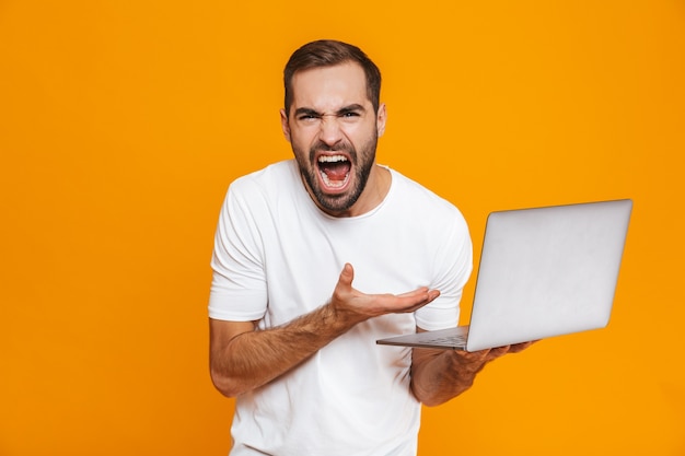 Portrait of irritated man 30s in white t-shirt screaming and holding silver laptop, isolated