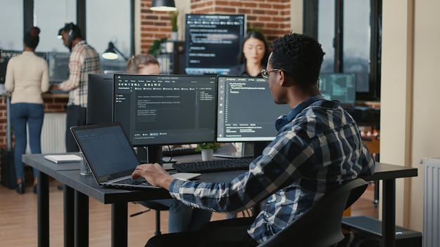 Portrait of system engineer fixing glasses while typing on laptop smiling satisfied about compiling results sitting at desk with multiple screens. App developer enjoying working on user interface.