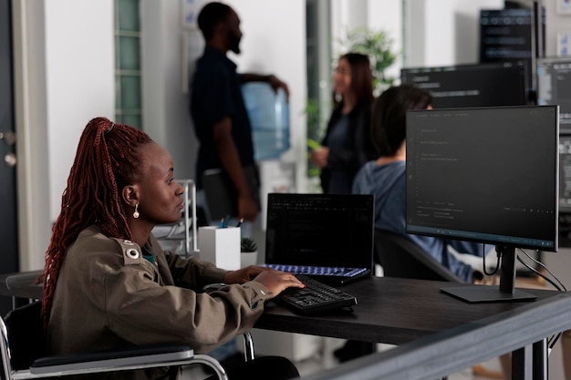 Programmer in wheelchiar typing html code on computer, working with cloud computing terminal window. Disabled software developer writing artificial intelligence algorithm in it company office.