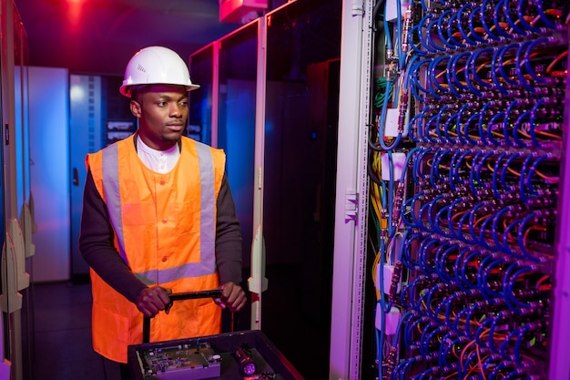 Serious black data center electrician in hardhat pushing cart with tools while coming server cabinet for repair