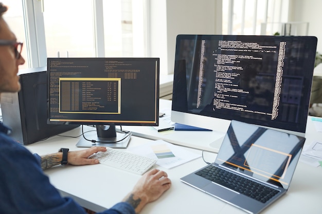 Side view at male programmer writing code while using multiple computers in IT developers office , copy space