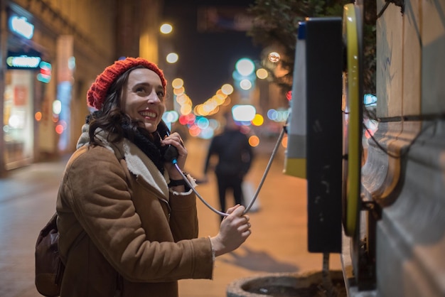 smiling woman talking at telephone booth while standing in city during night