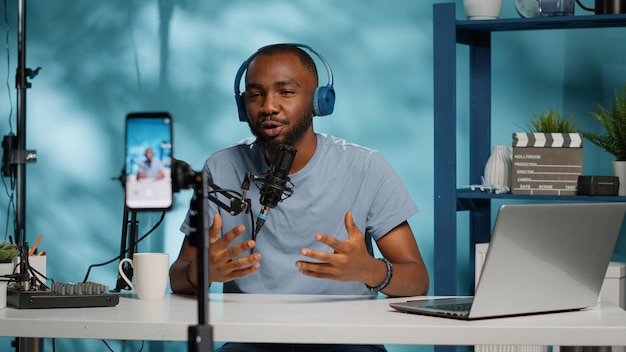 Social media influencer recording video for podcast channel using mobile phone on tripod. Blogger talking to subscribers, having microphone and headphones to film for vlog content.
