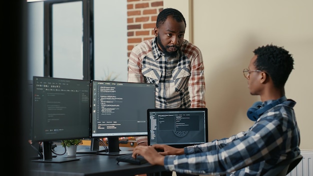 Software developer sitting at desk with multiple screens and laptop running code talking with colleague about artificial intelligence algorithm. Software developers doing teamwork in it office.
