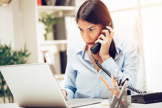 Successful young woman working at her desk in the office. She is using phone and working on laptop.