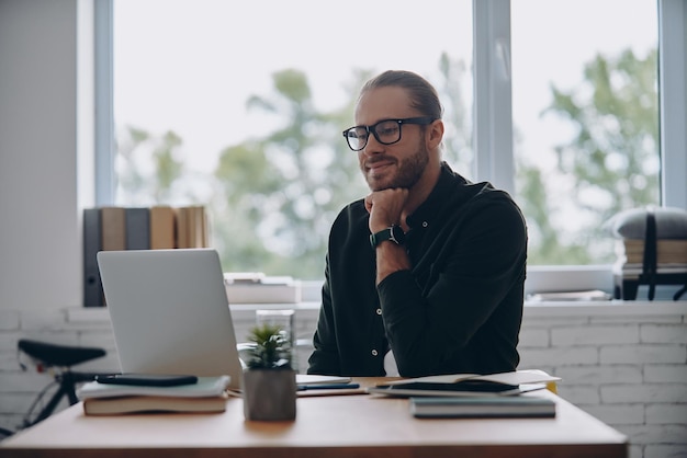 Thoughtful young man looking at laptop while sitting at his working place in office