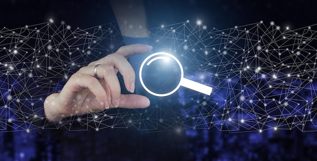 Tietojen hakutekniikan hakukoneoptimointi. Käsi pitää digitaalisen hologrammitietojen hakusymbolia kaupungin tummalla hämärtyneellä taustalla. Hakupalkki käyttöliittymälle. Hae osoitetta ja navigointipalkin kuvaketta.