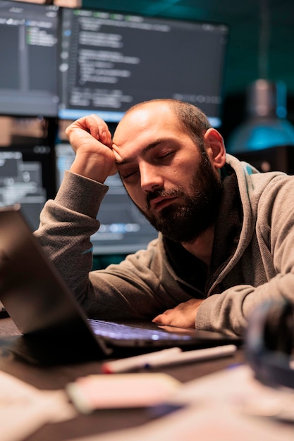 Tired IT enigneer falling asleep at office desk working on interface programming, holding head with hand late at night. Exhausted overworked developer sleeping after coding database system.