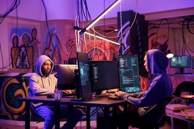 Two criminals breaching database to steal information and hacking server Asian and african american hackers in hoods programming internet virus and writing malicious software code on computer