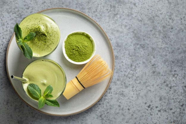 two glasses of dalgona matcha latte tea, bamboo whisk and matcha powder on grey. view from above. 