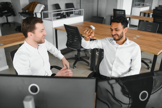Two men traders sitting at desk at office together monitoring stocks data candle charts on screen analyzing price flow smiling cheerful having profit teamwork concept