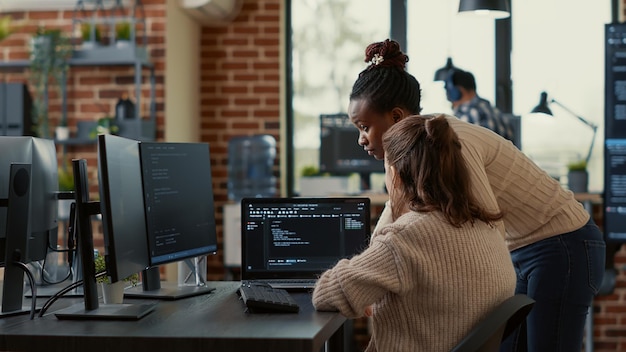Two system engineers analyzing source code on laptop looking for errors on screen while sitting at desk. Team of app developers working on group project for ai online cloud computing project.