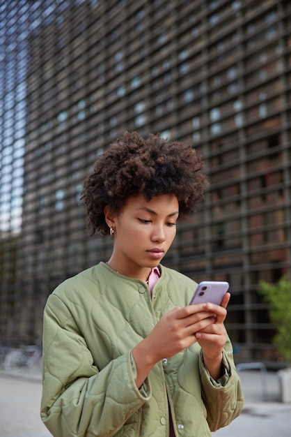 woman holds mobile phone for chatting online reads message on cellular checks mail dressed in jacket poses in urban place