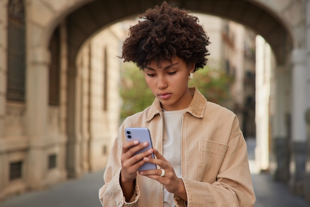 woman poses at urban setting with digital smartphone browses internet wears beige jacket stands outside has solo travel vacation