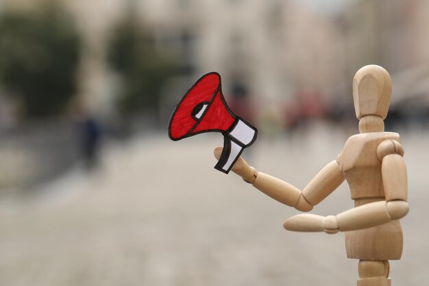 Wooden human figure with paper megaphone on city street closeup Space for text
