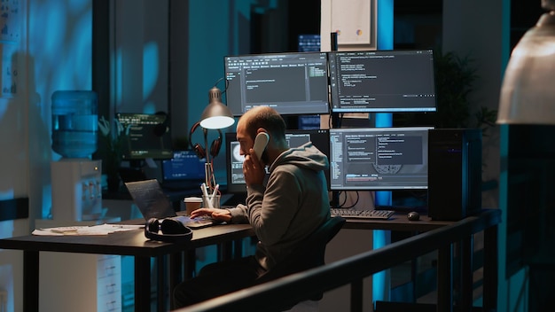 Young adult coder talking on landline phone at night, answering phone call in office. Male programmer using telephone with cord to chat and work on online cloud computing, artificial intelligence.