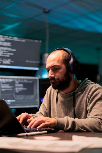 Young app developer writing code and listening to music on headphones, using artificial intelligence script to program big data on laptop. Programming server on terminal window.
