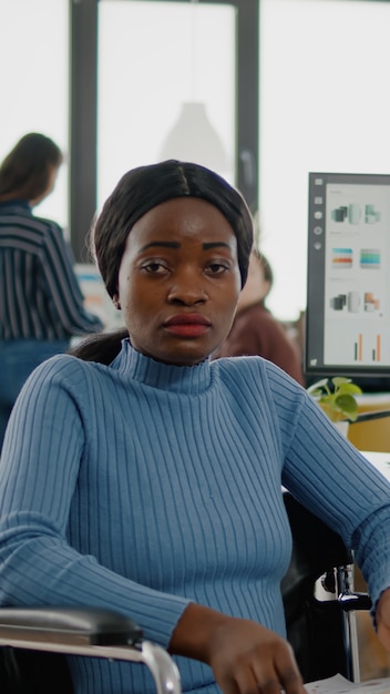 Young black woman with disabilities, invalid, disabled paralized handicapped looking at camera frustred, sad sitting in wheelchair in business office room, working at financial project with team.