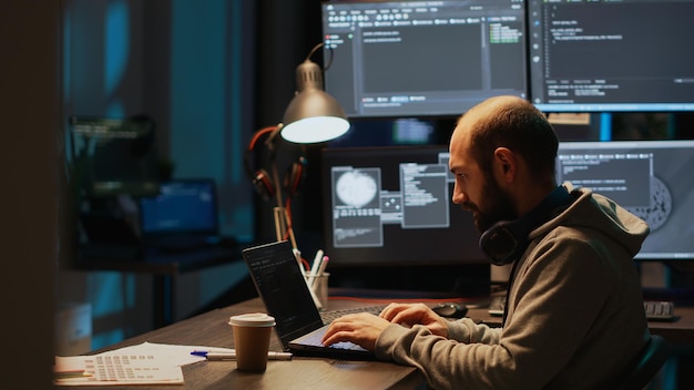 Young male developer typing server code on multiple screens using coding language on laptop late at night. Programming online firewall system on monitors to develop new html script.