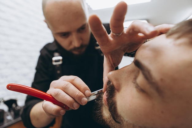 01062019 vinnitsa ukraine young brunette man with beard and\
bald barber holding hairdresser tools and haircut and modeling\
beard of client in modern barbershop