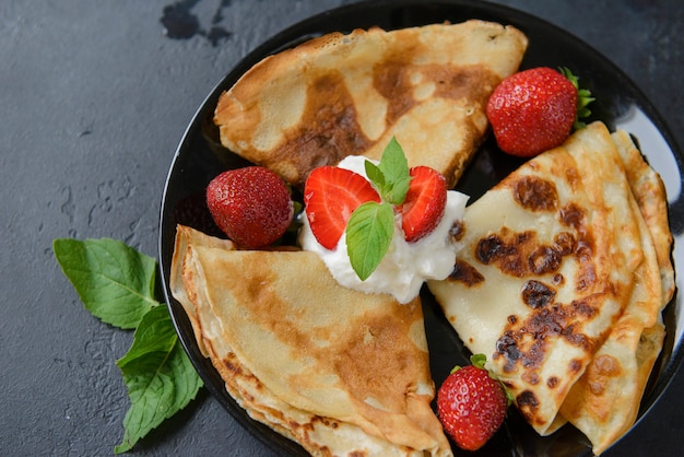 Photo 1 black plate with pancakes with strawberries and cream on a black surface, top view