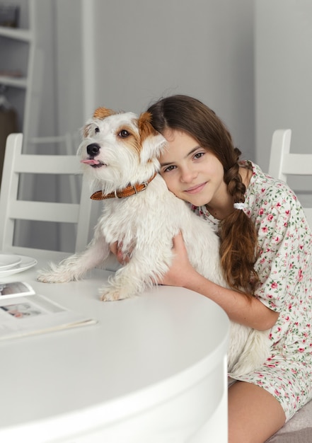 Foto 1 blank tienermeisje van 10 jaar zit aan de keukentafel en knuffelt hond jack russell's witbehaarde hond