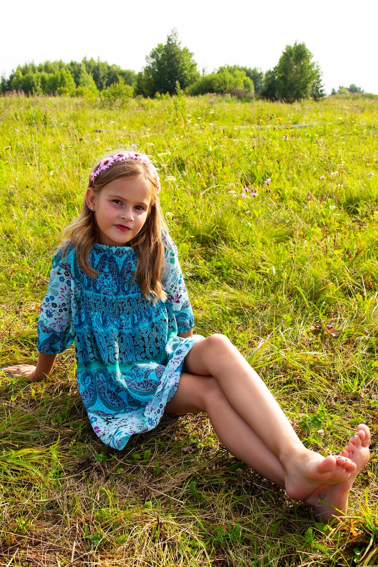 A 10-year-old girl in a blue dress sits on a field in grass and flowers, bare long legs, barefoot