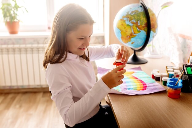 A 10-year-old girl draws a rainbow with paints at the table at home