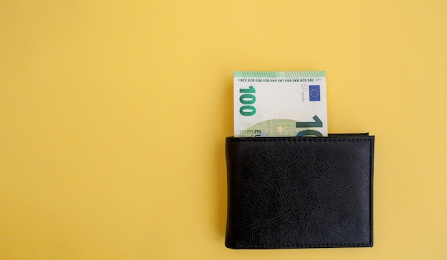 Банкнота в 100 евро торчит из черного кошелька на желтом фоне