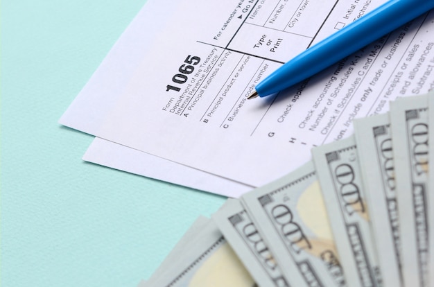 1065 tax form lies near hundred dollar bills and blue pen on a light blue background. 