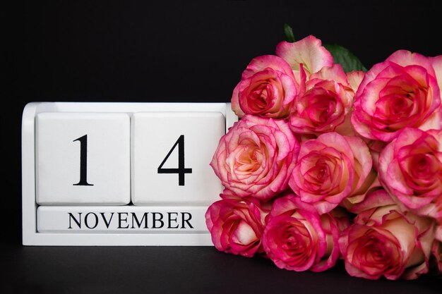 14 november houten kalender, wit op een zwarte achtergrond, roze rozen liggen in de buurt.