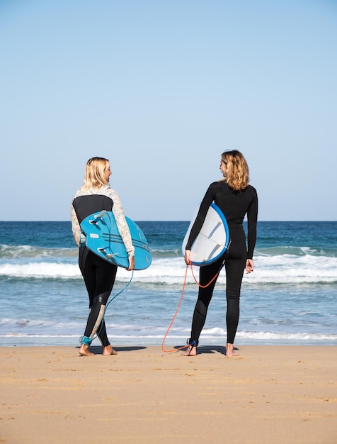 2 kaukasische surfervrouwen die op de strandkust staan en naar de golven kijken