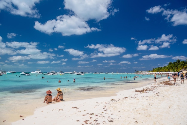 2 명의 섹시한 여자 숙녀들이 하얀 모래 해변 청록색 카리브 바다 Isla Mujeres 섬 카리브해 칸쿤 유카탄 멕시코에 브라질 문자열 비키니에 앉아 있습니다