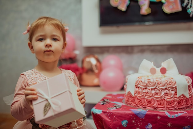 彼女の最初の誕生日ケーキとピンクのドレスを着た2歳の女の赤ちゃんお誕生日おめでとうcardaかわいい女の子は贈り物に囲まれて彼女の最初の誕生日を祝います