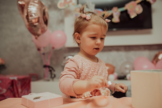 2 year baby girl in pink dress with her first birthday cake happy birthday carda cute little girl celebrates her first birthday surrounded by gifts