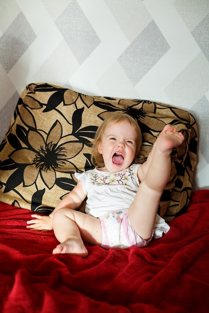 A 2 years old girl is sitting on the bed and smiling. The child rejoices and turns over. Happy child