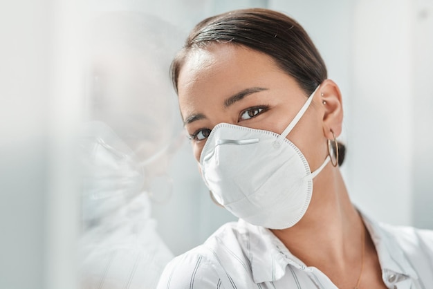 2020 The year of the face mask Shot of a masked young businesswoman leaning against a window in a modern office