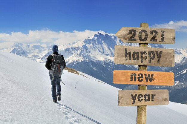 2021 happy new year written on a signpost with a hiker walking on the snow in a mountain