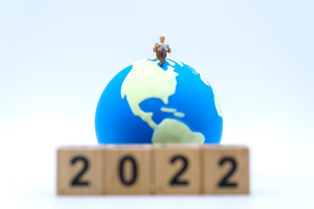 2022 New Year and Business Concept Closeup of businessman miniature figures sitting and reading a book or newspaper on mini world ball on white background with wooden number block