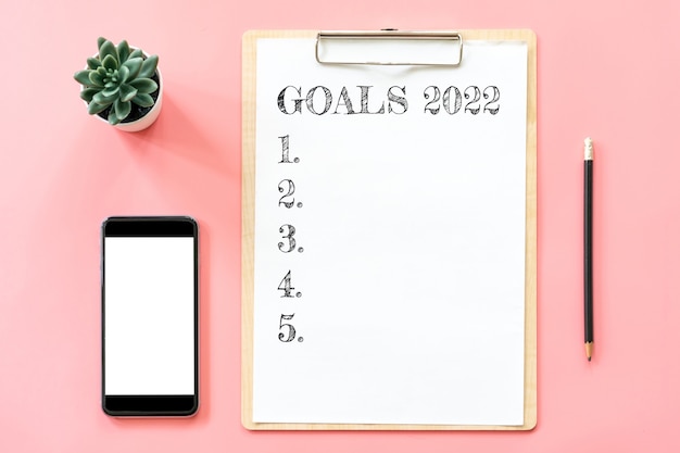 Photo 2022 new year concept. goals list in stationery, blank clipboard, smartphone, pot plant on pink pastel color with copy space