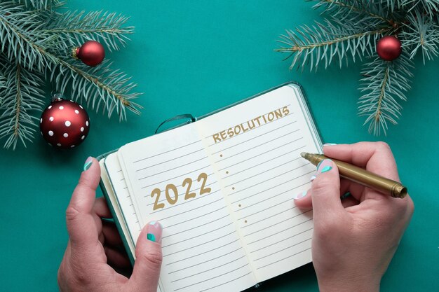 2022 New Year Resolutions concept flat lay Hands writing list in notebook Christmas background top view on fir twigs decorated with red glass baubles Xmas toys