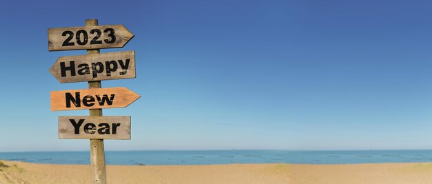 2023 happy new year written on a direction sign in front of a beach on blue sky